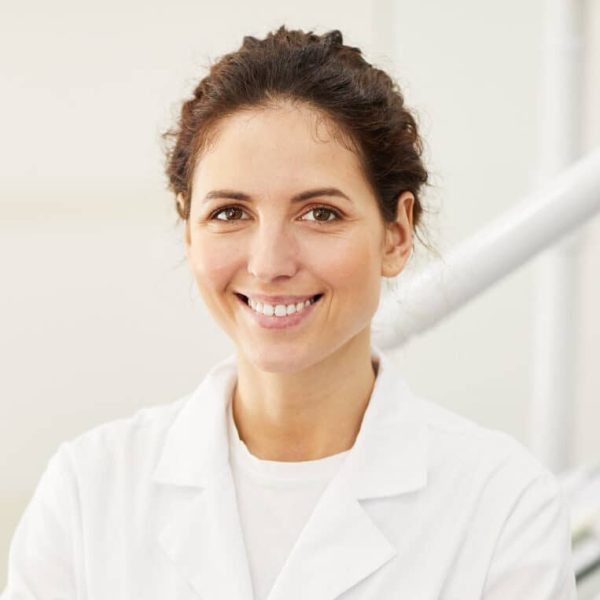 Waist up portrait of female dentist smiling at camera while consulting patient in background, copy space